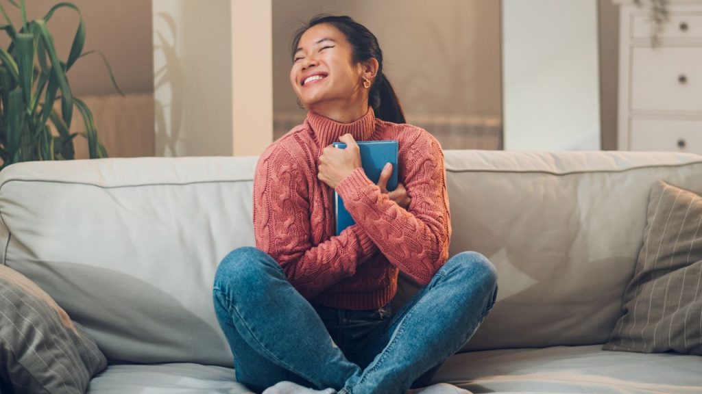 A girl, with a big smile, on a couch hugging a blue book.