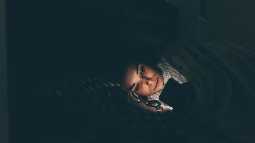 light of a phone emanating onto a woman's face in the dark bedroom.