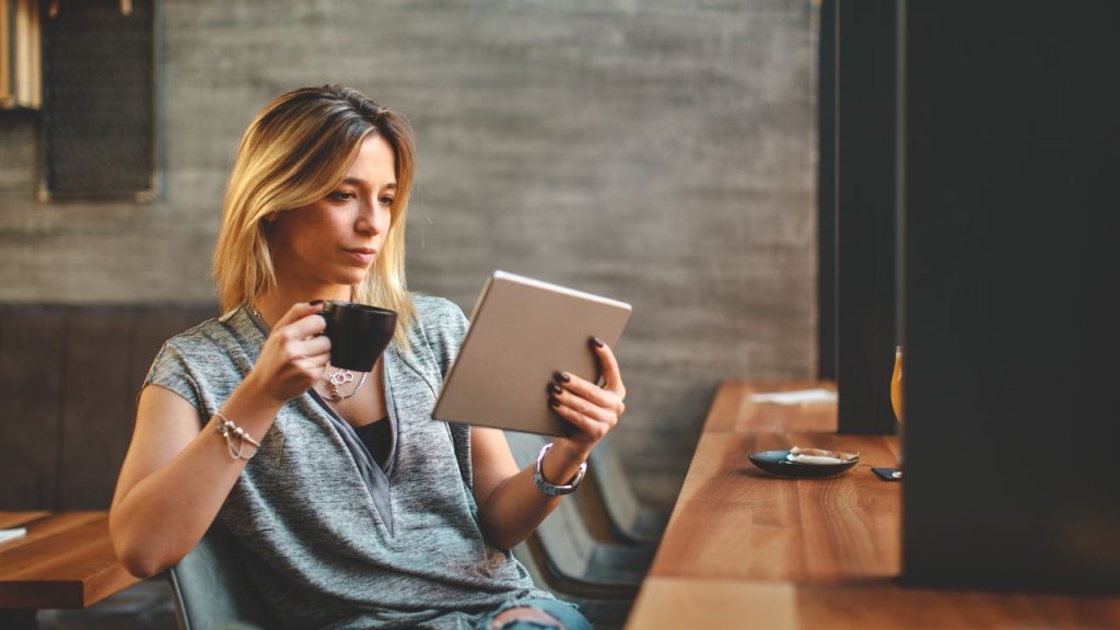 A woman sipping coffee in a coffee shop and reading off her electronic device.