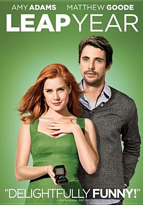 Red haired woman in green dress holding a ring with a brunette man standing behind her in a grey sweater