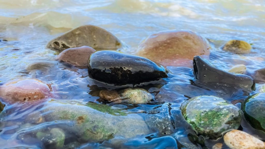 Rocks in a flowing river bed. The rocks are natural shades of black, red, green, and white. The water is a bit cloudy with sediments and organic matter.