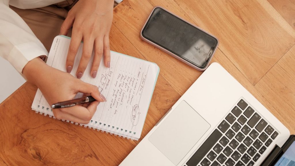 Person sitting at desk with laptop, phone and notepad. She is taking notes on the notepad.