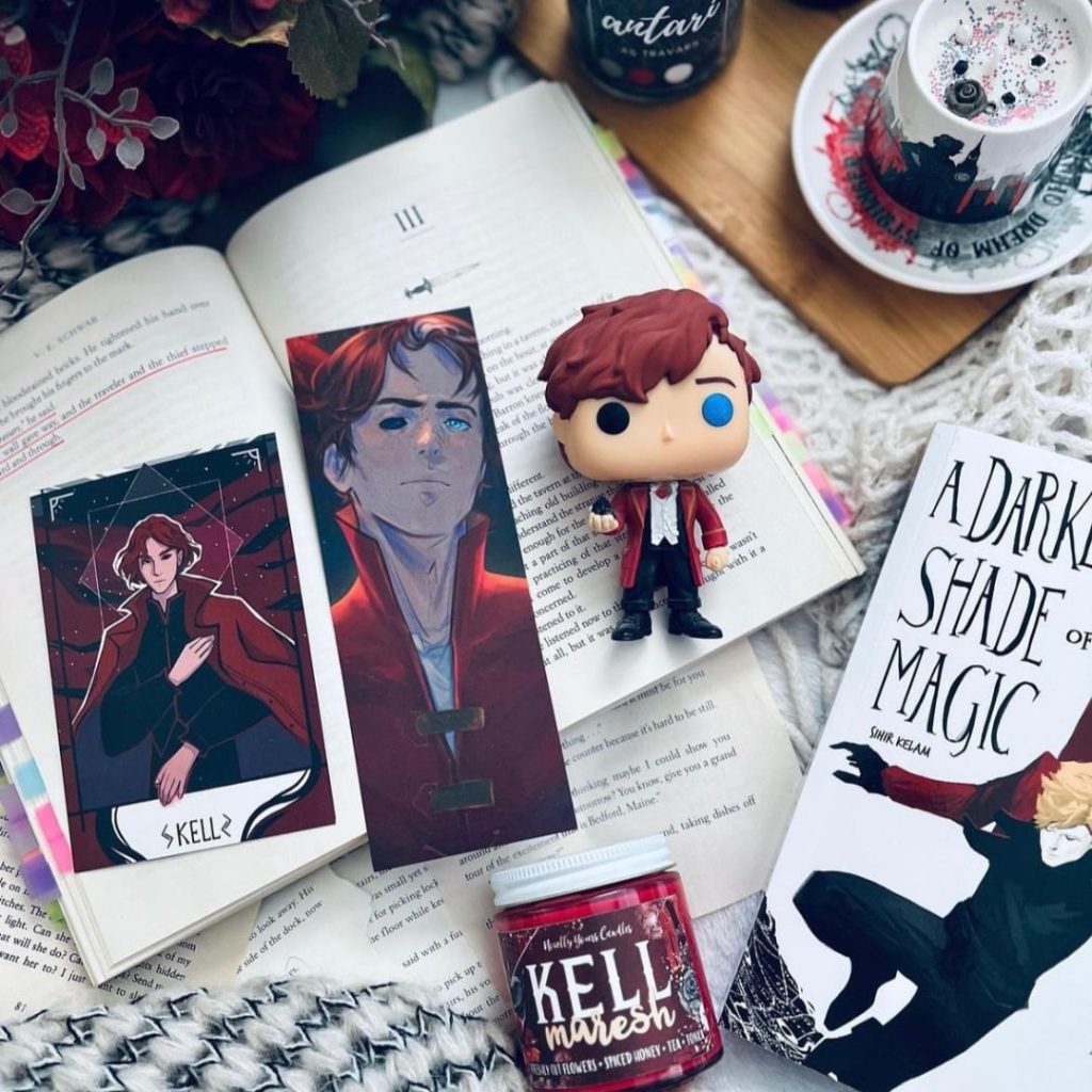 A merch spread of Shades of Magic character Kell is displayed. There is a candle, Funko Pop, art work and a bookmark that showcases Kell.