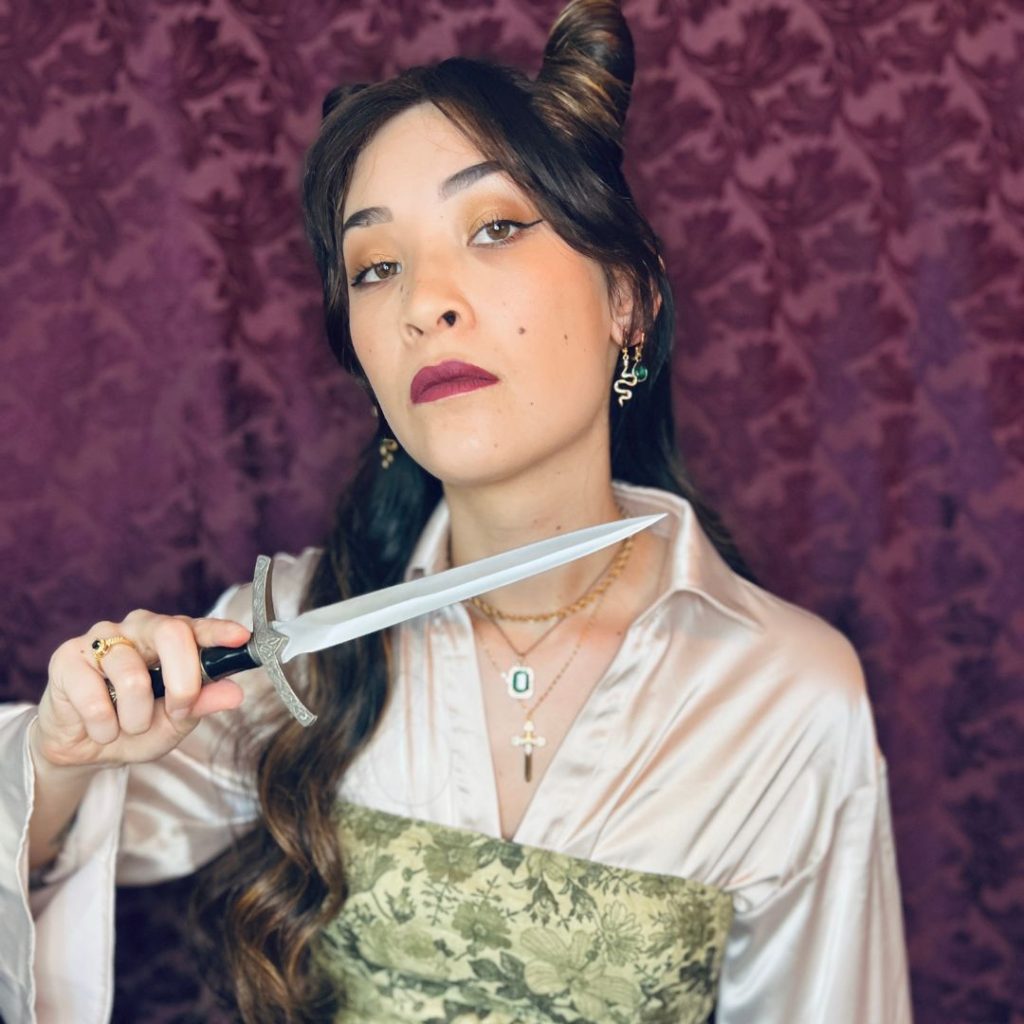 Kaylee stands in front of a mauve background as she holds a small dagger.