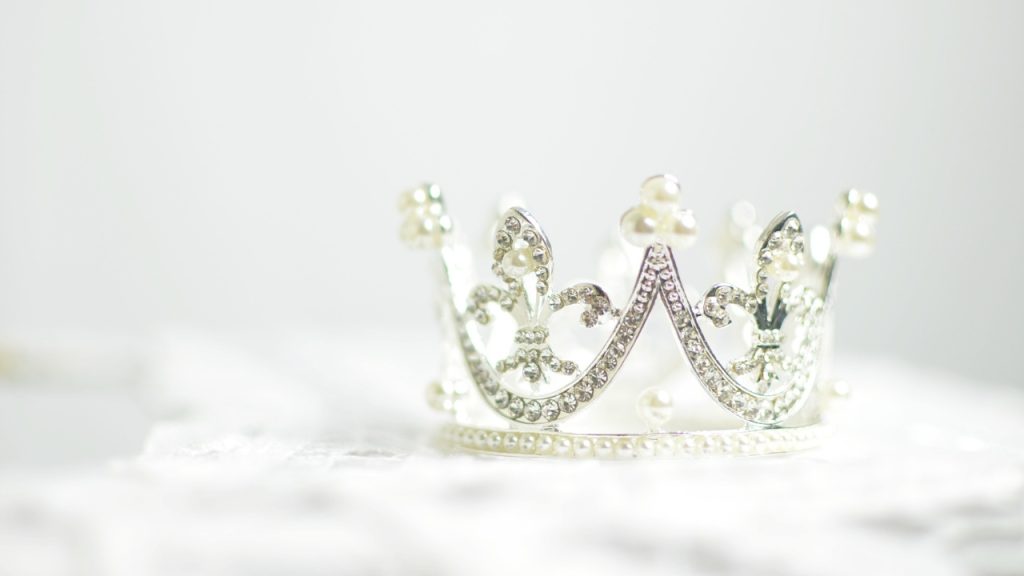A pearl and crystal encrusted silver crown placed on a textured white surface and against a white background.