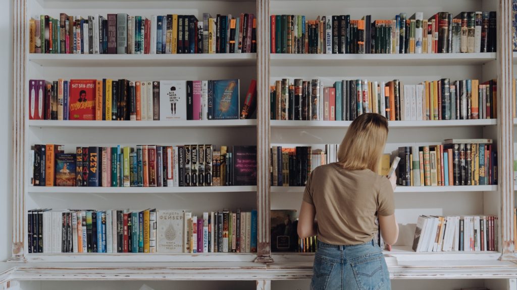 A set of white rustic bookshelves with a blond woman in a beige t-shirt and blue jeans storing a book into the shelves.