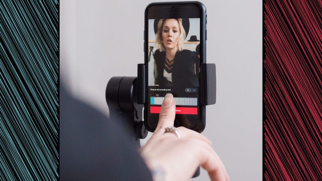 A woman with blonde hair, a black hat, and a black blazer presses record in the TikTok app. She has a rung on her finger, and her phone is held up by a tripod. The TikTok brand colors, blue and pink, border the image of the woman in diagonal lines.