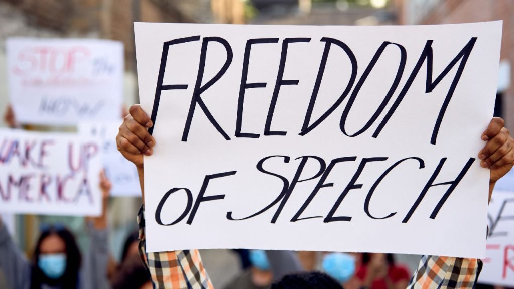 At a protest, a man in an orange, black, an white flannel holds up a paper sign with the words "Freedom of Speech" written in thick black marker and in all capital letters.