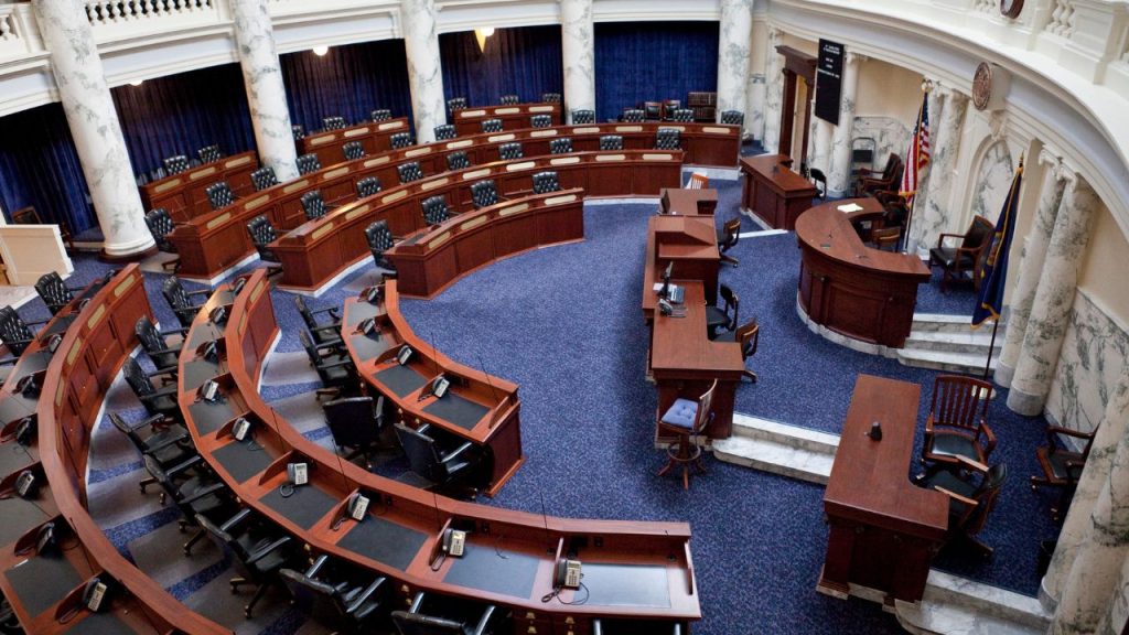 An aerial image of the House of Representatives floor. In a circular room, dark brown desks are arranged in semicircles on a blue carpet, towards a larger, straighter desk and a judge's desk. An American flag is at the front of the room, and white marble pillars span from the floor to ceiling.