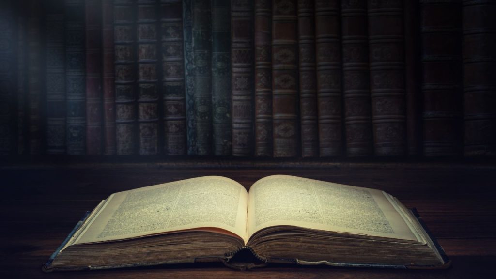 An open book with light shining on it in front of a darkened bookshelf.