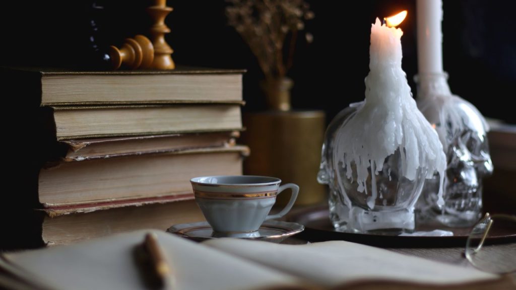 A desk featuring a stack of books, a cup of tea, and two candles with clear skulls as the base.