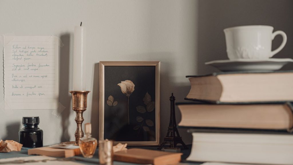 A desk featuring with a stack of books with a cup of tea on top. A slim candle in the background next to a framed picture of a flower.