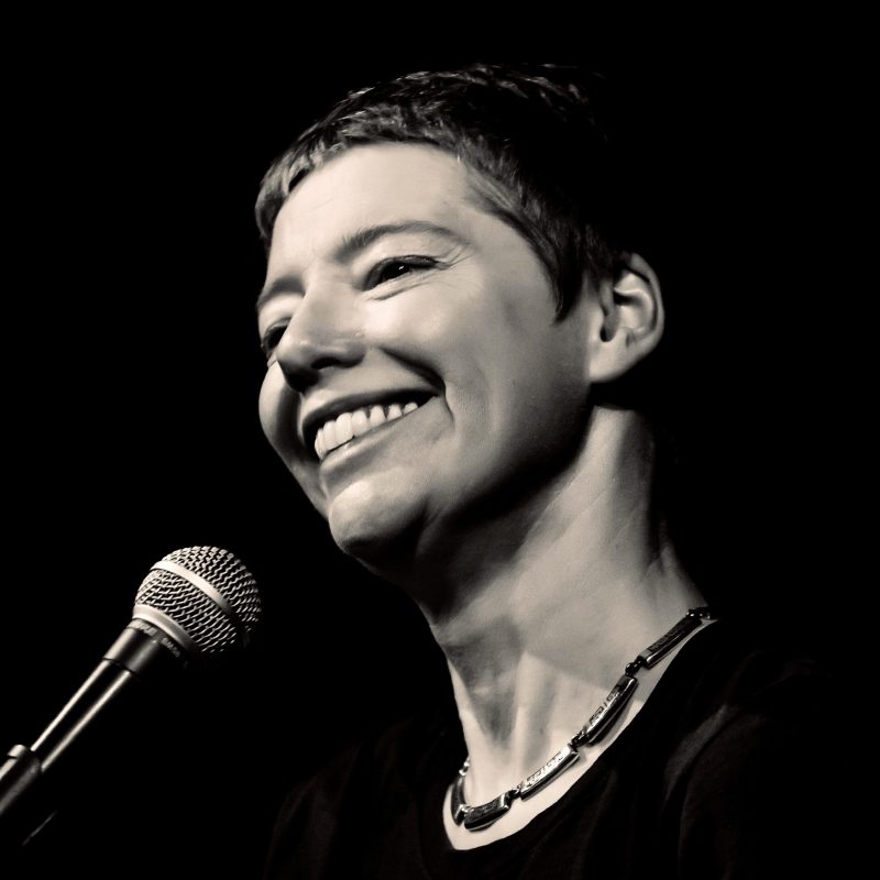 A black and whitephoto of Nicola Griffith smiling while standing in front of a microphone.