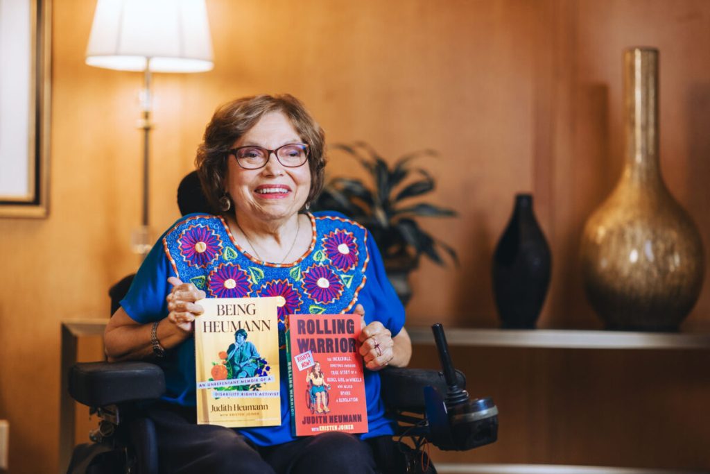 Judith Heumann smiling while sitting in her wheelchair and holding up her books 'Being Heumann' and 'Rolling Warrior.'