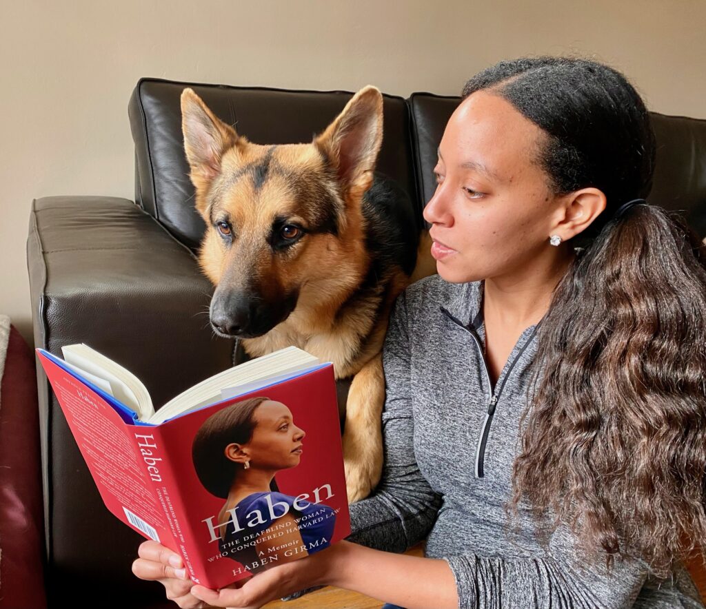 Haben Girma holding her book 'Haben: The Deafblind Woman Who Conquered Harvard Law' and sitting with her seeing eye dog Mylo.