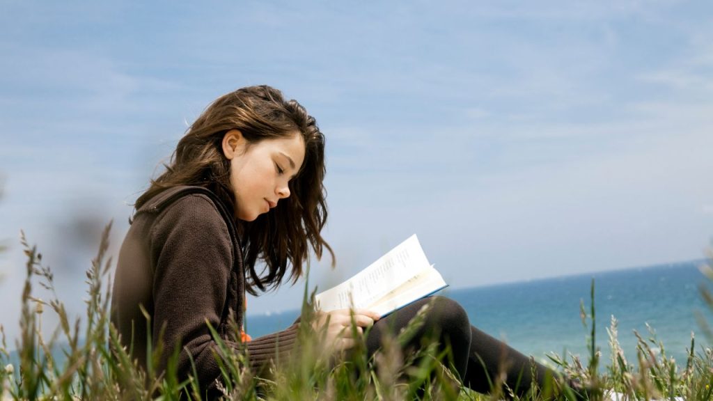 A girl reading a book in a field with a body of water behind her.