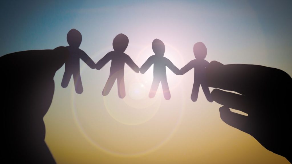Two hands hold out a connected cut out of four paper people to the sun so that the hands and paper are solid black and the edges of the sky are blue.