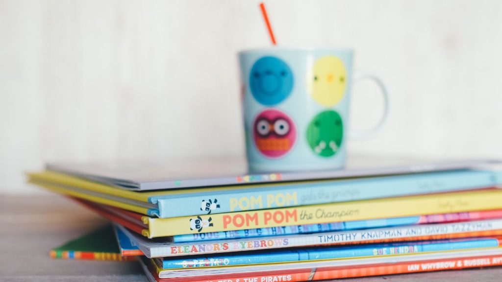 A mug with colorful and silly circular cartoon faces holds a bright orange straw on top of a stack of colorful children's books. Everything is on a wooden tabletop against a white background.