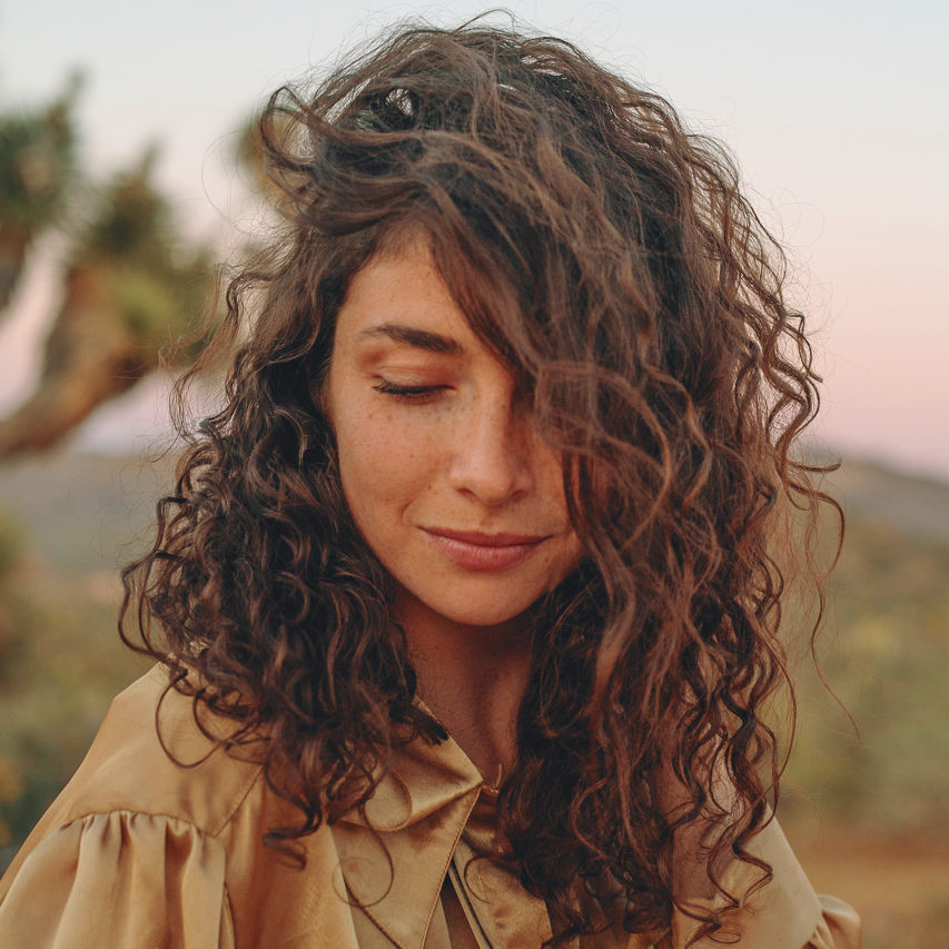 A portrait of Madison Margolin. Her brown curly hair is swept to one side, partially covering her face as she smiles and looks down. She is outside in a desert environment with the sun setting in the background.