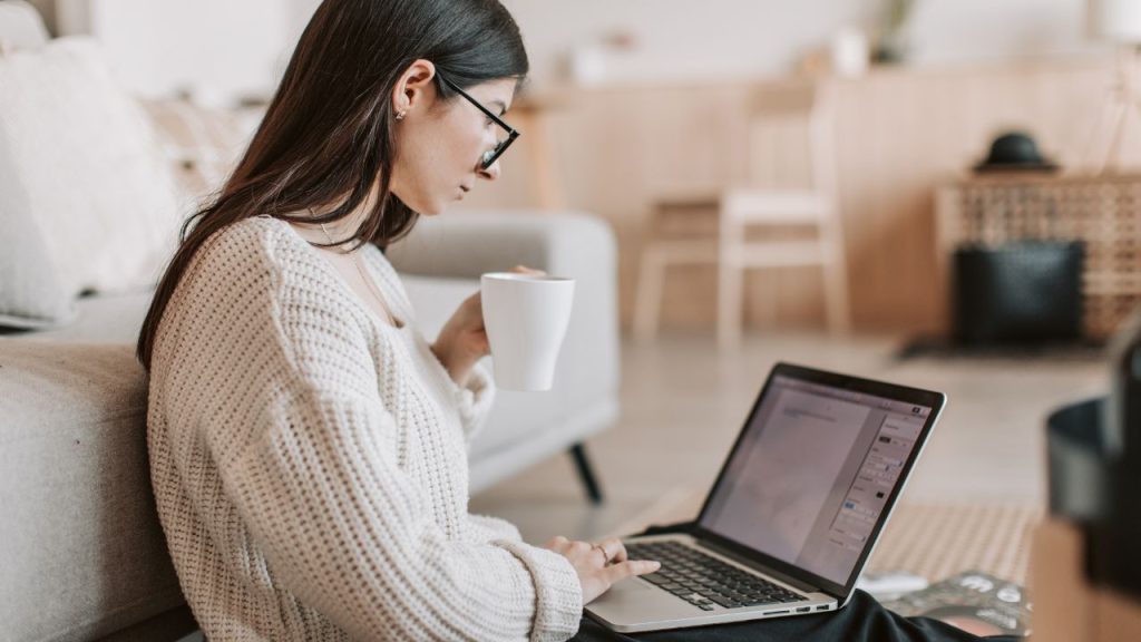 A woman with long, straight, dark brown hair scrolls in a blank document on a laptop. In the other hand she holds a white mug. She is sitting on the floor in front of a gray couch in a white sweater, and is in a minimalistic room.