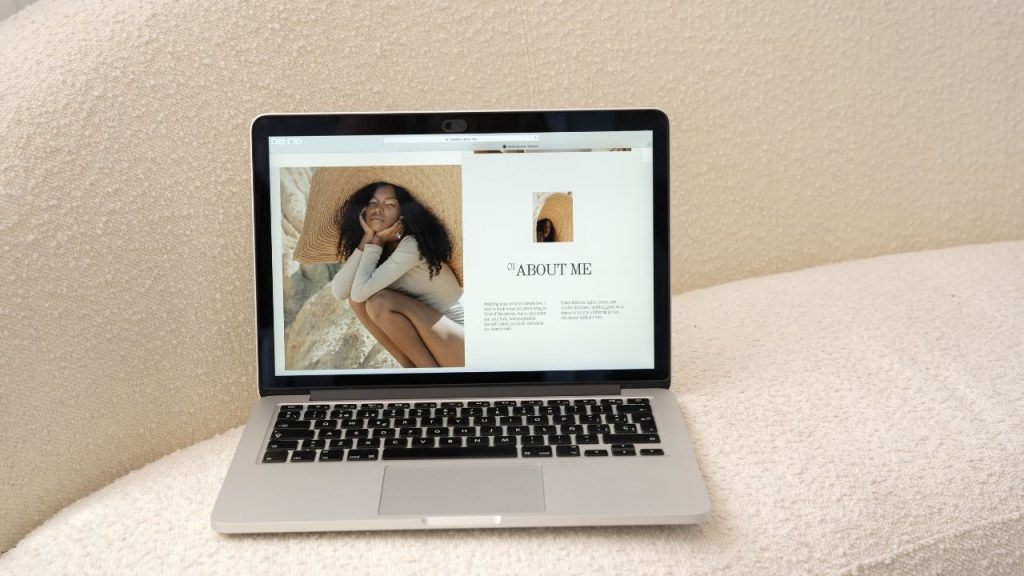 On a laptop on a beige couch is a woman's portfolio. The words "About Me" and two paragraphs are on one side of the screen, and next to it is a large portrait of a woman with black curly hair, a cream-colored long sleeve and matching shorts, and a large, floppy straw hat.