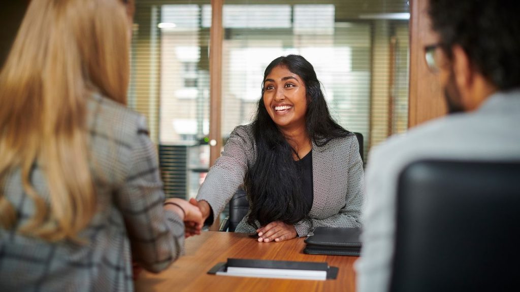 In an office, three people sit at a desk. One person is facing the photo, and the other two are have their back to the viewers. The single person, a woman, shakes the hand of another woman on the opposite side. Everyone is wearing professional office attire at this interview.
