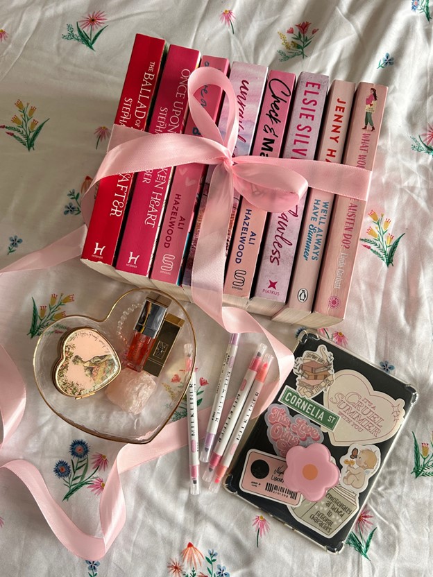 In a Bookstagrammer collage, a series of romance books in pink covers are tied together with a silky pink bow. In front of are a heart shaped glass bowl with a vintage heart-shaped trinket box, pink and gold lip-gloss, a pink crystal, and pearl bracelet. Besides the bowl are pink and purple highlighters, as well as a tablet with pink and heart-shaped stickers. Ribbon is intertwined between all of the objects, and everything is on dainty floral bedsheets.