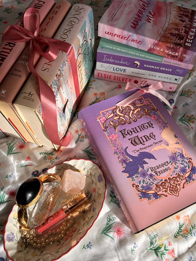 On dainty floral bedsheets, two stacks of colorful books frame another, single book laid in the forefront. One book stack is tied with a dark pink ribbon. Fourth Wing by Rebecca Yarros is centered with a tiny, light pink ribbon on its top right corner. Besides it is a vintage floral, porcelain dish with a black gem and gold bracelet, makeup with gold accents, and a pink crystal.