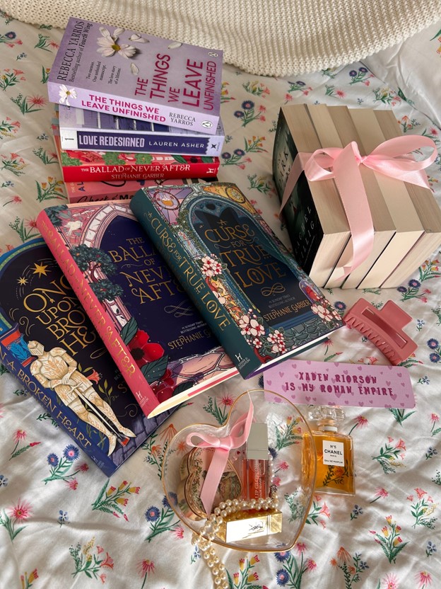 Three stacks of books are posed on floral bed sheets. Three books in a series are spread in the forefront. The books stack to the right are held together by a pink ribbon. In front of all the books is a heart-shaped glass bowl with a pink lip-gloss, a heart-shaped, vintage trinket box, and a string of pearls. Chanel perfume lies on the side of it, as well as a pink bookmark with tiny hearts on it, and a pink claw clip.