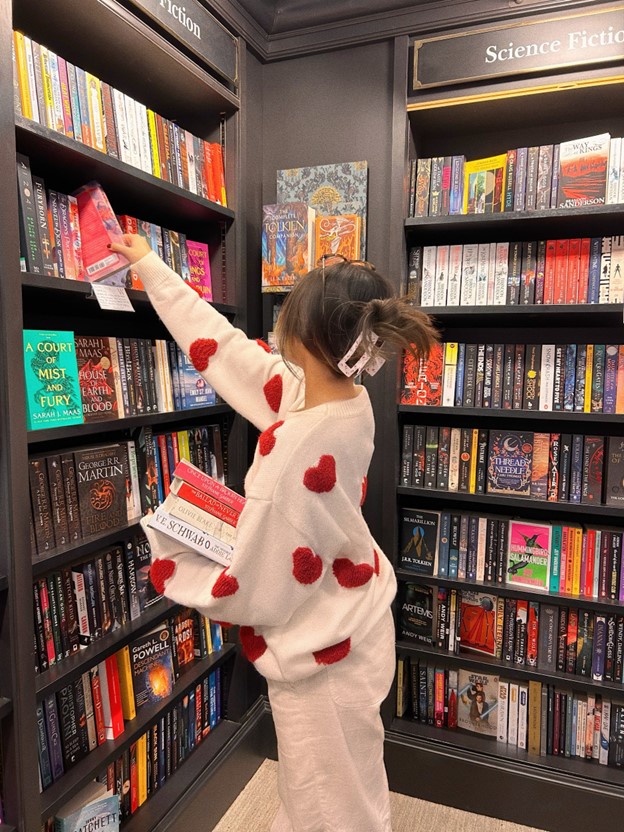 Bookstagrammer reaches for a book in the fantasy section of a bookstore. The bookshelves are made of dark wood. She holds a stack of four books in her hand and is wearing a pink sweater with fuzzy red hearts.