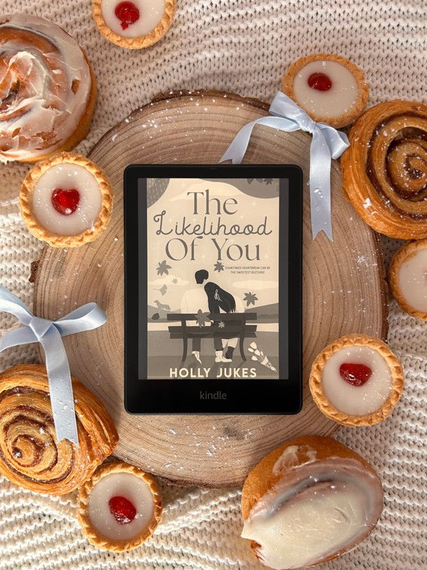 The book jacket for The Likelihood of You by Holly Jukes is presented on a Kindle. The Kindle rests on a wooden stump on a fabric background. Tarts with red jelly and other pastries frame the Kindle.
