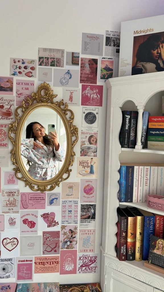 Image of Bookstagrammer Andrea posing in a royal-style wall mirror. Around the mirror, small posters of various art prints, musicians, and album colors. They are all in shades of pink. Besides it is a white bookshelf with books stacked neatly inside of it.