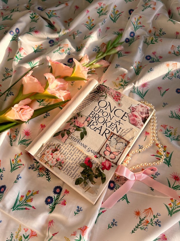 Once Upon a Broken Heart by Stephanie Garber is placed on floral bedsheets with the cover pulled back. Pink floral and butterfly stickers are collaged on the title page. Around the book are fresh pink flowers, a string of pearls, and a pink silk ribbon.