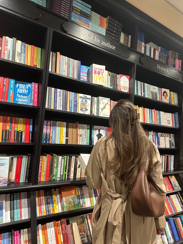 Bookstagrammer Andrea poses in front of dark black bookshelves in a bookstore. Her back is turned to the camera as she reads the back of a book.