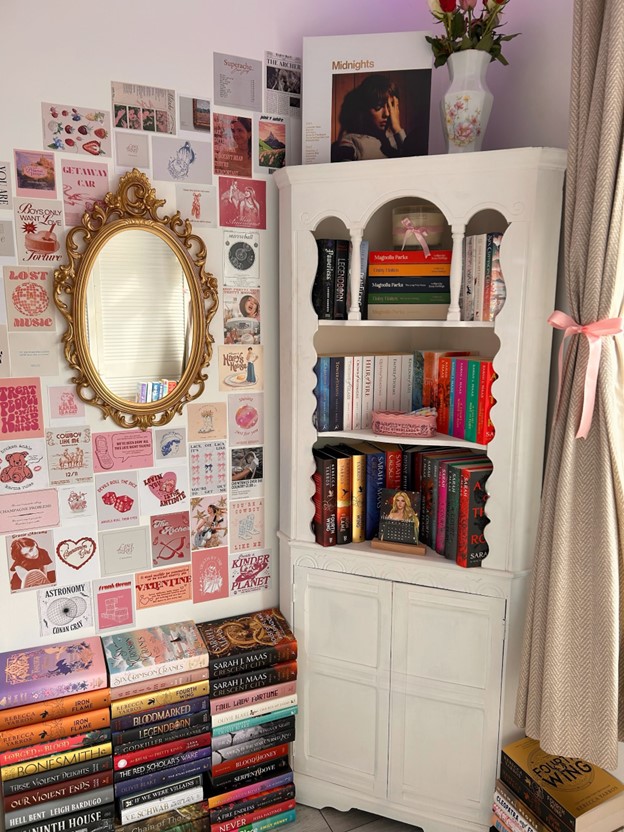 A brightly lit room with the curtains pulled back with a pink ribbon, a white bookshelf neatly stacked with colorful books, and a gold, ornate mirror with tiny square posters in shades of pink covering the wall around it. In front of the mirror are three pillars of books stacked on the floor.