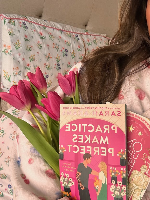 Bookstagrammer Andrea poses on her floral bed with pink tulips and two books with pink covers in one of her arms. The book in the front is Practice Makes Perfect by Sarah Adams.