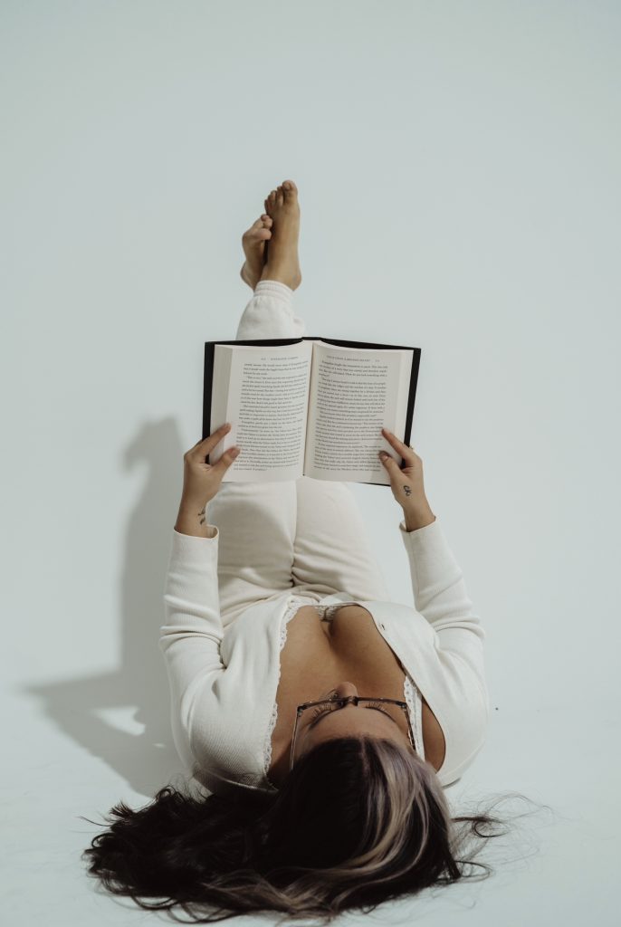 Photo of Bookstagrammer Cassandra of @romance.and.rose lying on her back on the ground of a white room. Her feet are up against the wall and she holds open a book with a black cover to read.
