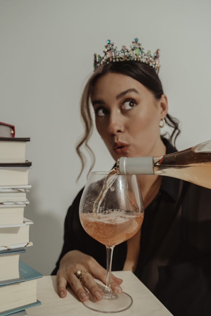 Photo of Bookstagrammer Cassandra of @romance.and.rose with her hair in a low ponytail and a bejeweled crown on her head. She pours rose into a wine glass. Besides her is a stack of books.