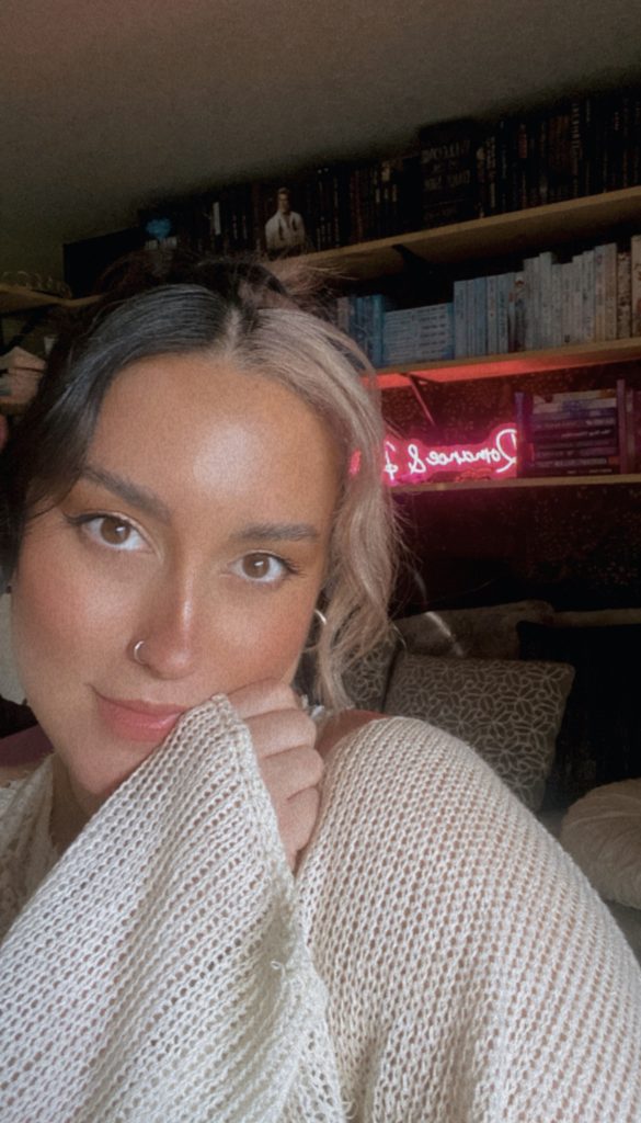 Photo of Bookstagrammer Cassandra of @romance.and.rose taking a selfie in front of her bookshelf. On the shelf is neon pink lighted sign. Cassandra is in a white sweater and poses with her head tilted and hand resting on her shoulder.