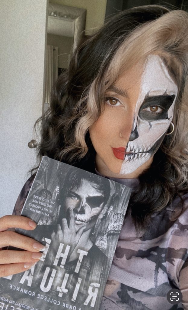 Photo of Bookstagrammer Cassandra of @romance.and.rose with skeleton face paint on one half of her face. She holds up a books with a man on the cover in matching face paint.