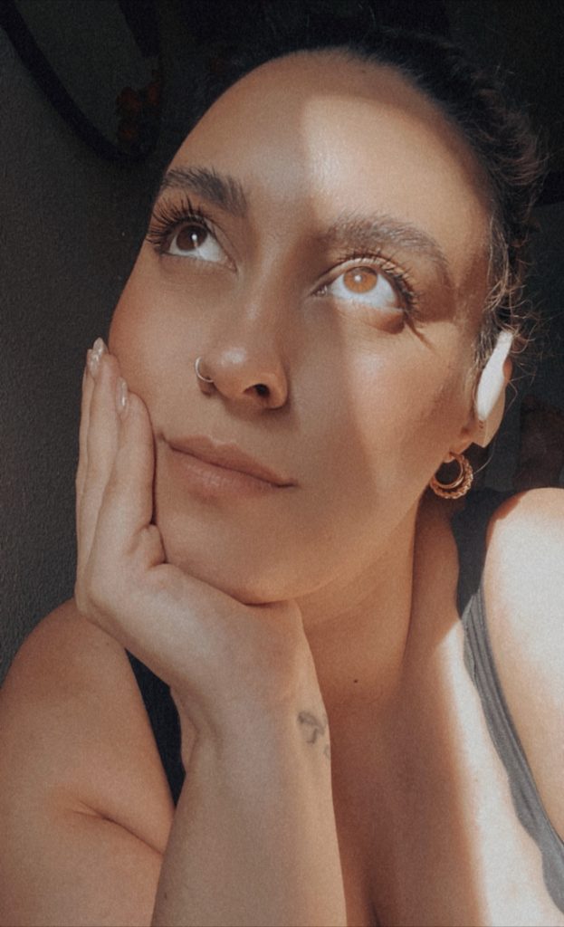 Photo of Bookstagrammer Cassandra of @romance.and.rose posing in the sunlight with her head resting in her hand. Her hair is up and she has a silver nose ring, two gold hoop earrings, and a black tank top.