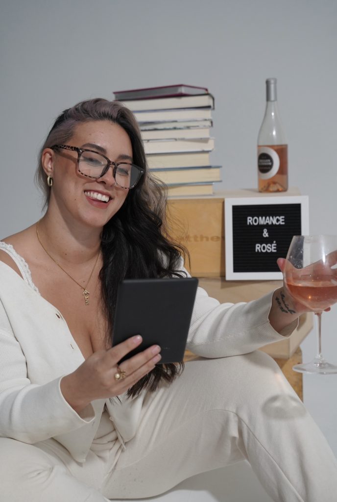 Photo of Bookstagrammer Cassandra of @romance.and.rose reading on her kindle in one hand and holding a glass of rose in another. Behind her are wooden crates with books stacked on top, as well as a bottle of rose.