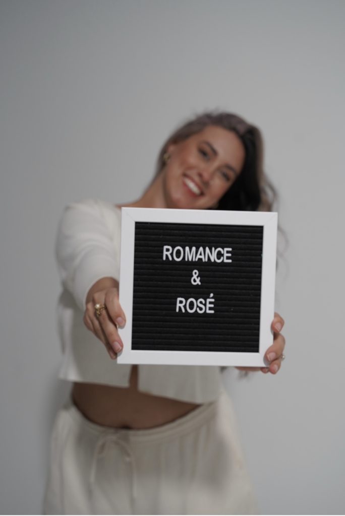 Photo of Bookstagrammer Cassandra of @romance.and.rose holding a black sign in a white frame with her Instagram name written in white letters. She is in a white loungewear set, posing in front of a white wall with her head tilted to the side.