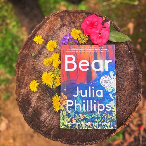 Bear by Julia Phillips placed on a large brown tree trunk. Dandelion flowers, along with three purple flowers and two pink, frame the book.