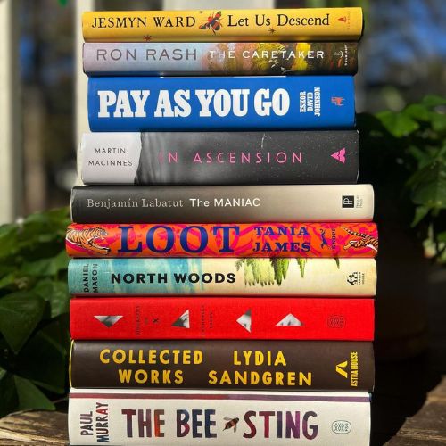 A stack of hardcover literary fiction books on a wooden bench outside. Blurry in the background are lush green shrubs and a blue sky.
