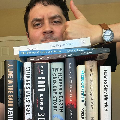 Photo of Bookstagrammer JC Grenn posing above a lined-up stack of books, with three extra books laying horizontally on top of the stack. He holds a thumbs up.