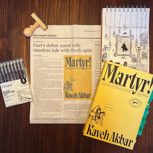 Bookstagram collage for the novel Martyr by Kaveh Akbar. The book itself, with a bright yellow cover and blue annotation tabs poking out of the pages, lies on top of a notebook with a doodle of a giraffe in an ornate but homely room. The left is a newspaper page of a book review for Martyr, a stamp, and a case of black drawing pens. Everything is on a dark hardwood surface.