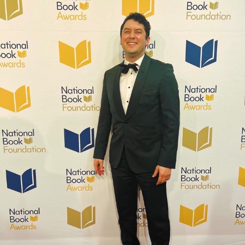 Photo of Bookstagrammer JC Grenn in a dark green suit in front of a wall with the National Book Awards logo.