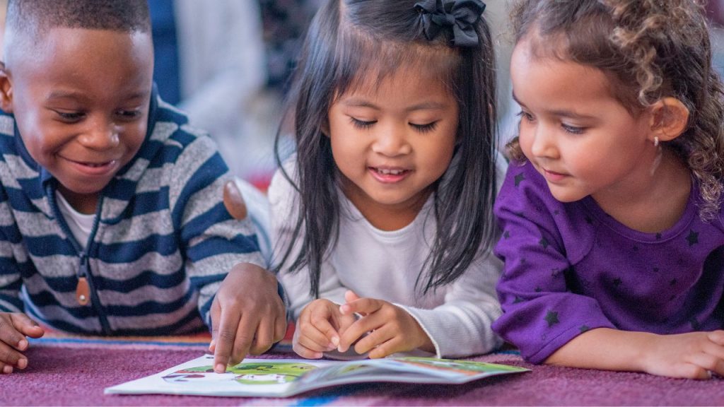 Three multicultural children lie on their stomachs on a purple carpet reading a picture book together.