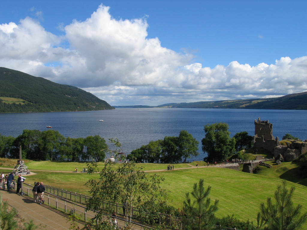 Loch Ness in Scotland, where greenery surrounds a large, blue lake. On one side are green-covered mountains. On the other side are trees and a town with people walking along a road beside the lake.